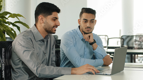 Two Indian employees sitting in modern office using laptop, young colleagues discussing project. Arab serious millennial male businessmen satisfied with result of negotiations concluded contract