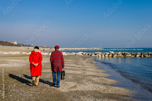 Due persone vestite di rosso camminano lungo la spiaggia del Lido di Venezia in una giornata invernale di sole photo