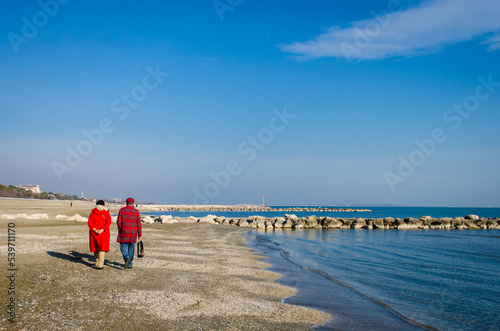 Due persone vestite di rosso camminano lungo la spiaggia del Lido di Venezia in una giornata invernale di sole photo