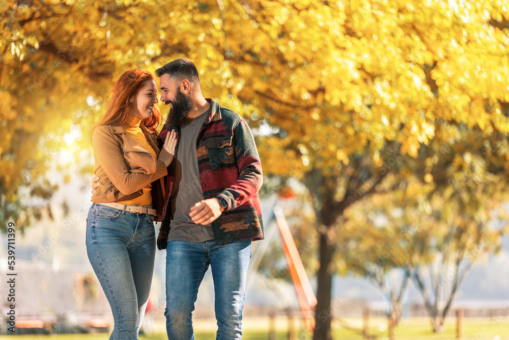 Fototapeta premium Young happy couple looking at each other while holding hands and walking in autumn park.