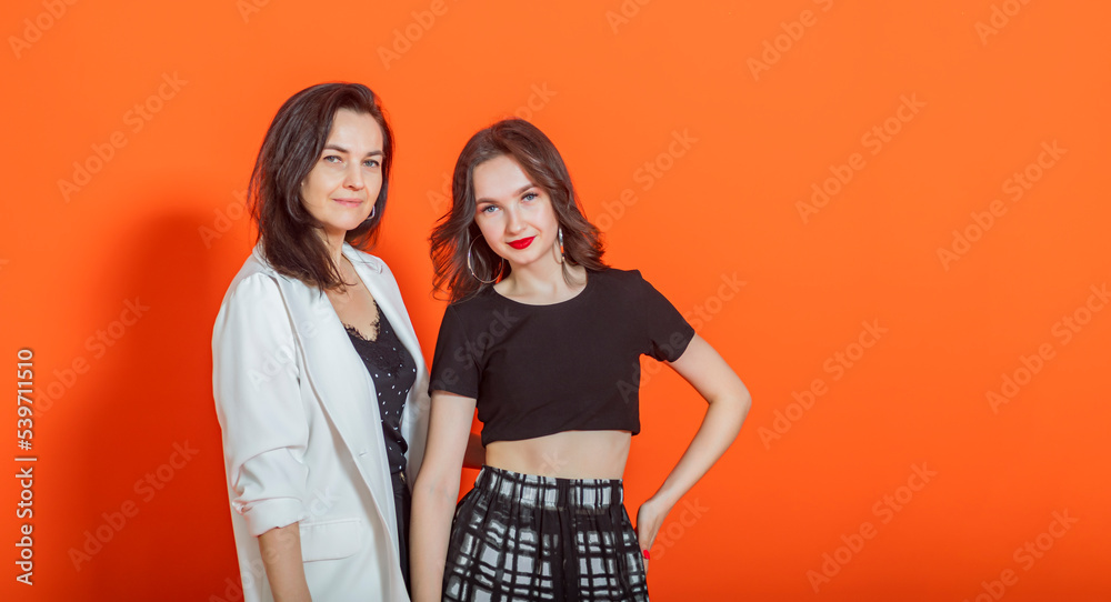 Two young brunette girls (family - mom and daughter), in casual clothes, stand on orange background, smile and look at camera. Two young brunette girls (family - mom and daughter), in casual clothes, stand on orange background, smile and look at camera.