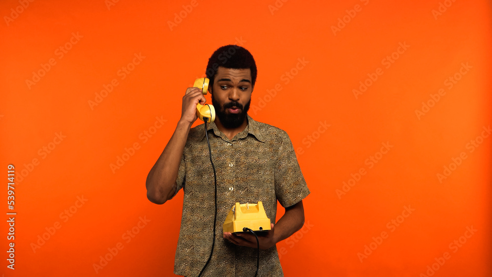 surprised african american man with beard talking on retro telephone on orange background.