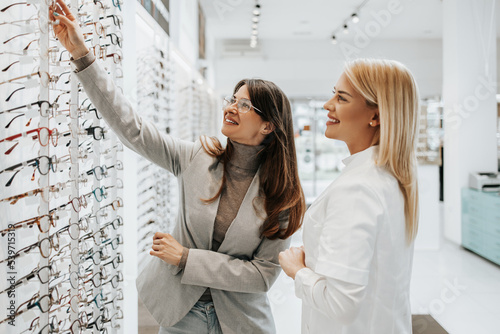 Beautiful and fashionable woman choosing eyeglasses frame in modern optical store. Female seller specialist helps her to make right decision.