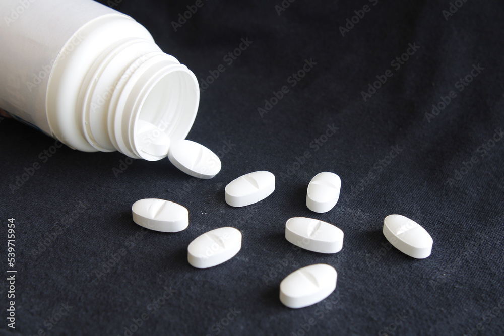 Botella de plàstico blanca con medicamento en pastillas para tratamientos de enfermedades volcadas sobre la mesa, forma un original diseño farmacèutico con fondo negro Botella de plàstico blanca con medicamento en pastillas para tratamientos de enfermedades volcadas sobre la mesa, forma un original diseño farmacèutico con fondo negro
