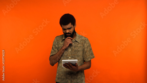 thoughtful african american young man with beard holding digital tablet on orange background.