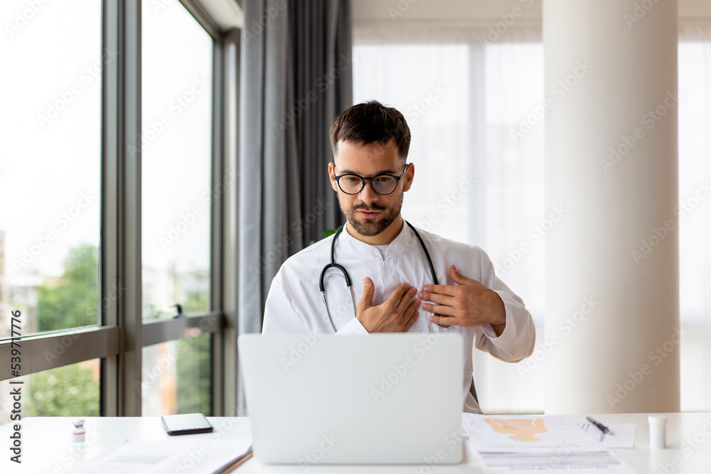 Fototapeta premium Concentrated young physician working on computer looking at laptop screen thinking on problem solution concept. Serious man doctor wearing white coat doing online research at workplace.