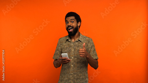 excited african american man using smartphone and showing like on orange background.