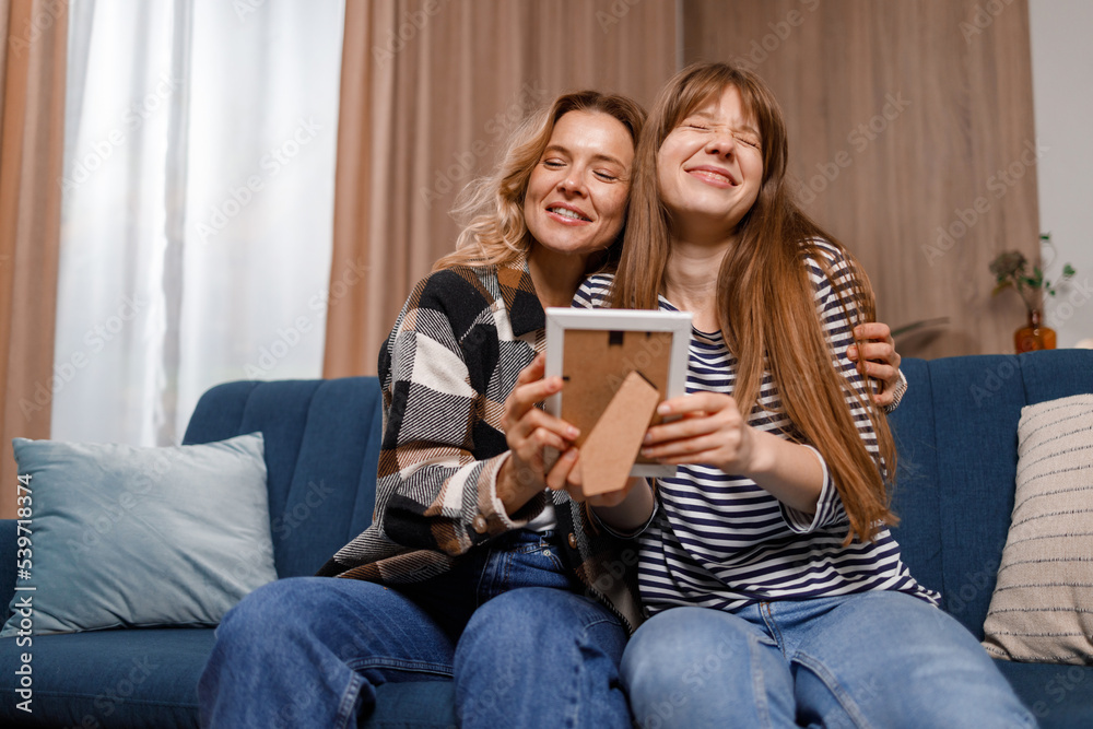 Mother and daughter sitting on the couch in the living room hugging each other looking at an old photo in a frame Mother and daughter sitting on the couch in the living room hugging each other looking at an old photo in a frame