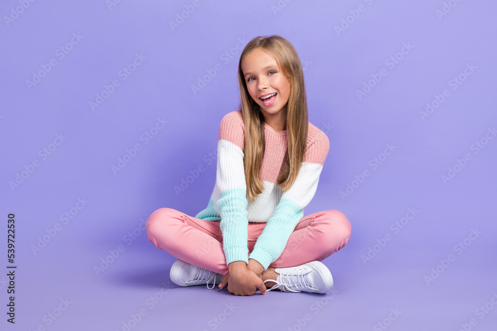 Full length photo of cute nice satisfied girl toothy smile legs crossed sitting floor empty space isolated on purple color background Full length photo of cute nice satisfied girl toothy smile legs crossed sitting floor empty space isolated on purple color background