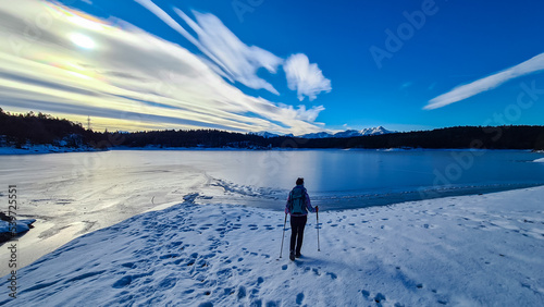 Rear view of woman with hiking backpack watching the frozen lake Forstsee, Techelsberg, Carinthia (Kaernten), Austria, Europe. Winter wonderland at sunrise. Snow capped mountain Mittagskogel (Kepa) photo