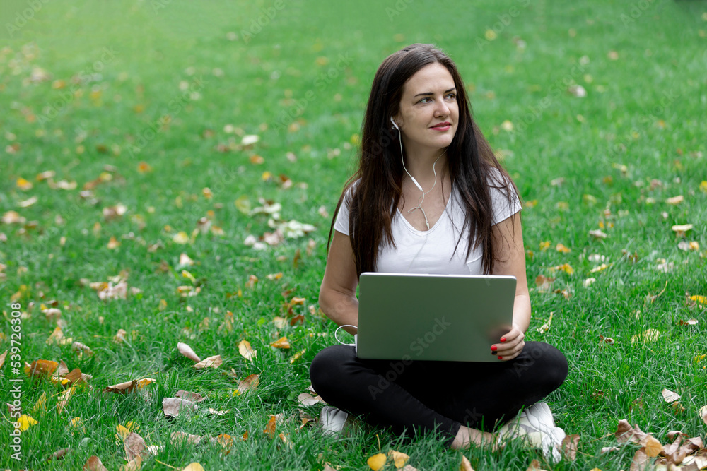 Portrait of a pretty young woman sitting on the green grass in the park on a summer day with a laptop.