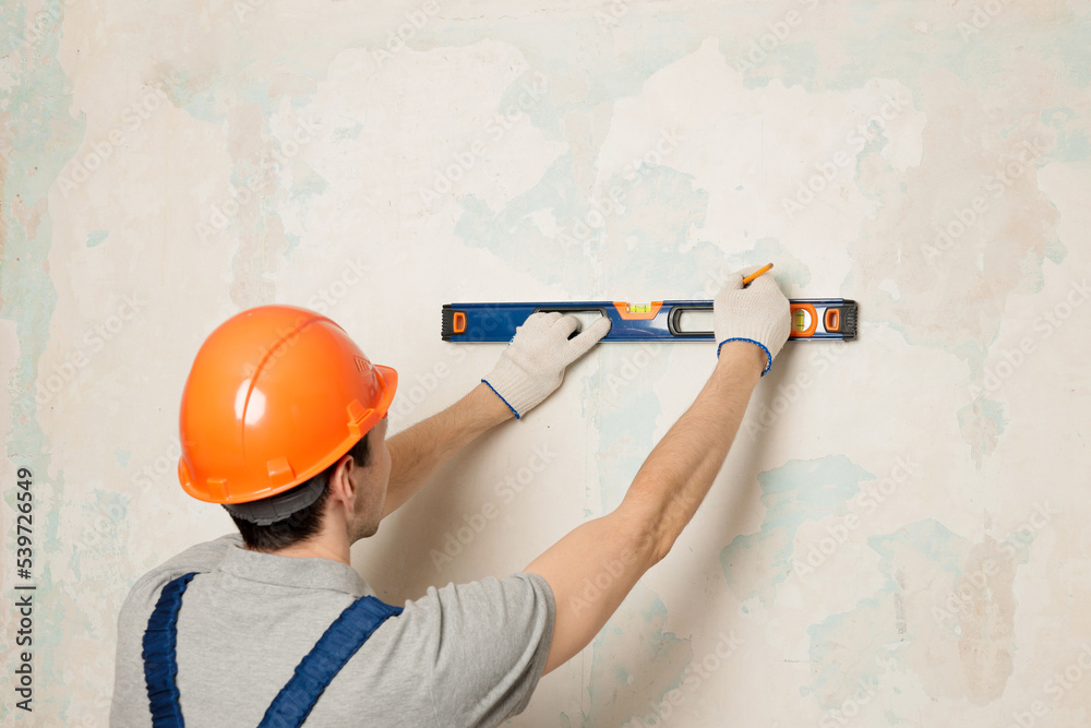 Handyman, builder in uniform measuring the wall using bubble level. Repairman in blue overalls working with a bubble level, home renovation and construction concept. Copy space. Handyman, builder in uniform measuring the wall using bubble level. Repairman in blue overalls working with a bubble level, home renovation and construction concept. Copy space.