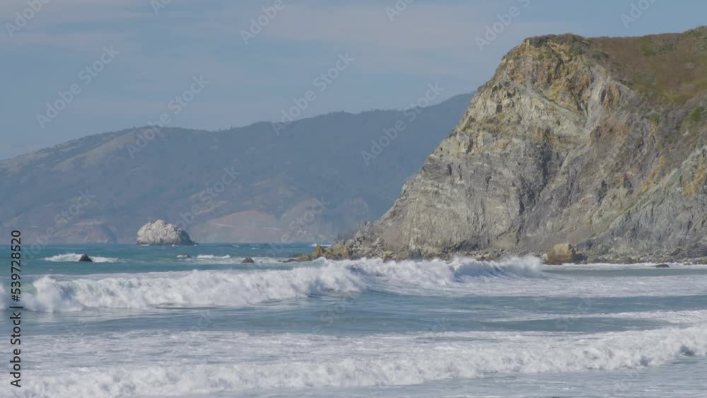 Stationary shot of waves crashing into the side of a cliff with mountains on the background located in Big Sur California Beach. Stationary shot of waves crashing into the side of a cliff with mountains on the background located in Big Sur California Beach.