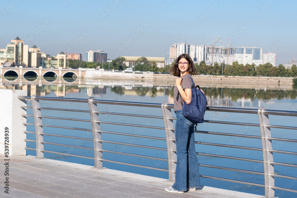 Smiling young curly-haired college student dressed in a T-shirt and jeans holding a backpack standing on the city river embankment, full height. The concept of student travel Smiling young curly-haired college student dressed in a T-shirt and jeans holding a backpack standing on the city river embankment, full height. The concept of student travel