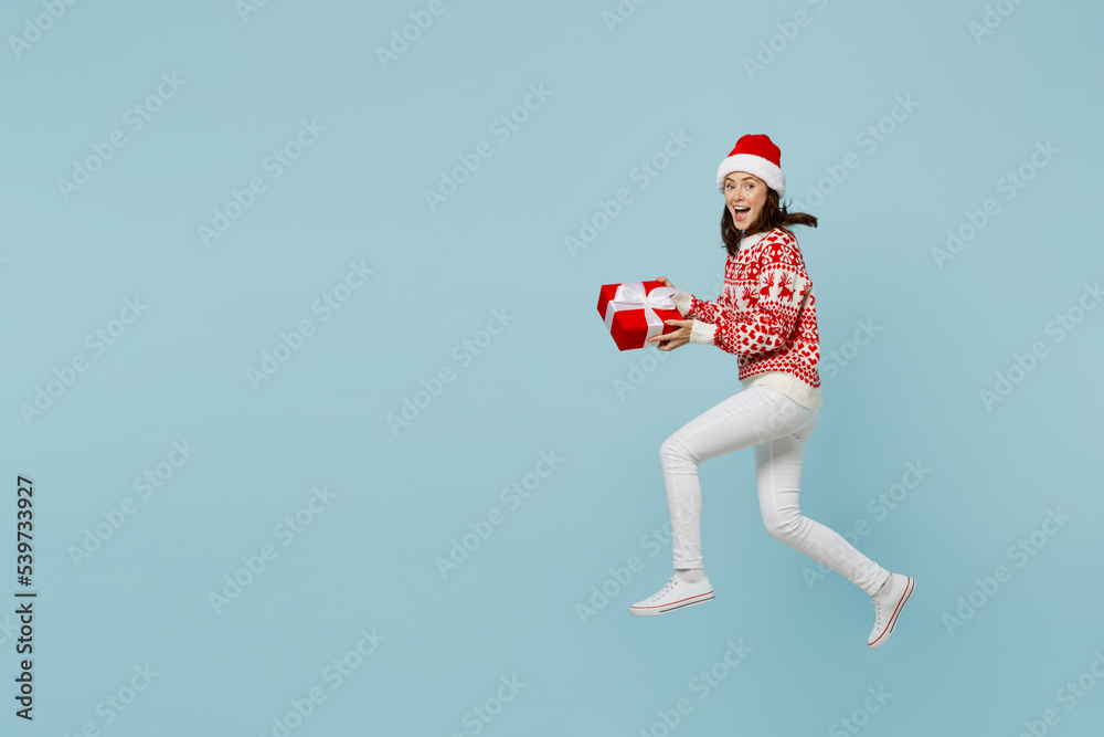 Full body young merry woman in red sweater Santa hat posing jump high run hold present box with gift ribbon bow isolated on plain pastel light blue cyan background Happy New Year 2023 holiday concept. Full body young merry woman in red sweater Santa hat posing jump high run hold present box with gift ribbon bow isolated on plain pastel light blue cyan background Happy New Year 2023 holiday concept.