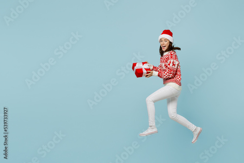 Full body young merry woman in red sweater Santa hat posing jump high run hold present box with gift ribbon bow isolated on plain pastel light blue cyan background Happy New Year 2023 holiday concept.