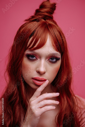  Beautiful young woman with red hair and bright pink makeup on a pink background. Woman touching her lips with her hand
