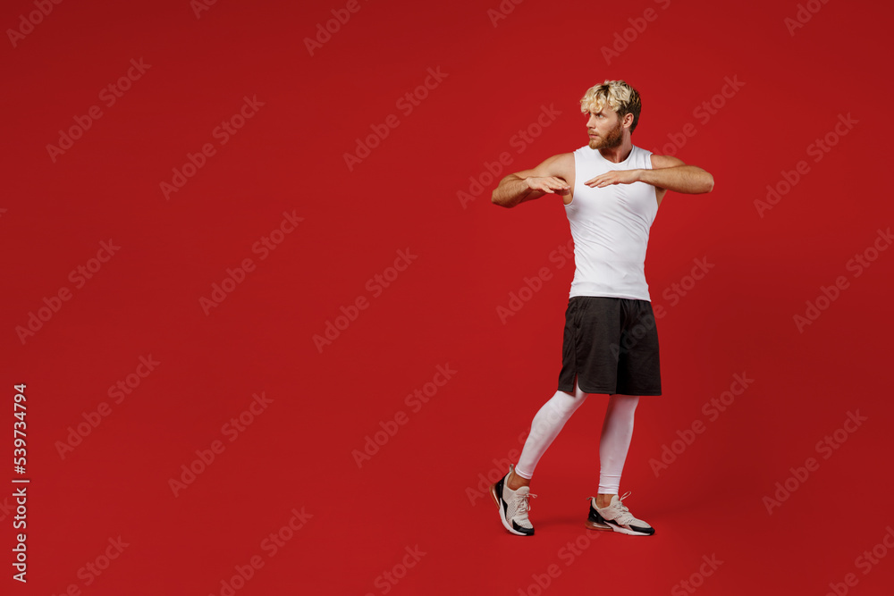 Full body sideways young strong sporty toned sportsman man in white clothes spend time in home gym do hand exercise look aside on area isolated on plain red background Workout sport fit body concept