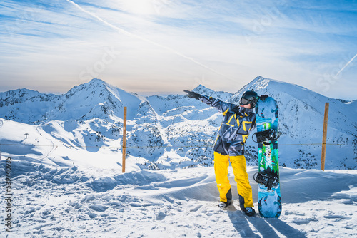 Portrait of a snowboarder with a snowboard showing with hand distant piste on the top of the snowy Pyrenees Mountains. El Tarter, Grandvalira, Andorra