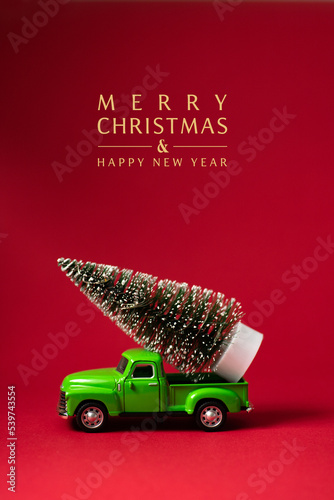 Green retro toy car carrying a Christmas tree on red background with text - Merry Christmas and Happy New Year. Christmas and New Year celebration concept. Greeting card. Copy space, selective focus