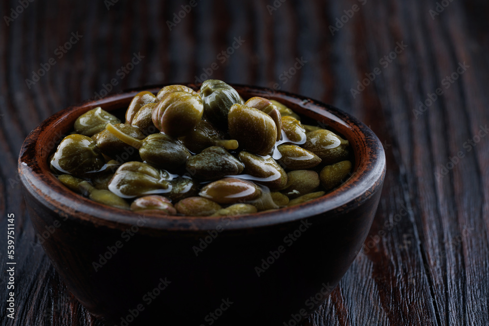 An earthenware bowl filled to the brim with capers stands on a wooden table. An earthenware bowl filled to the brim with capers stands on a wooden table.
