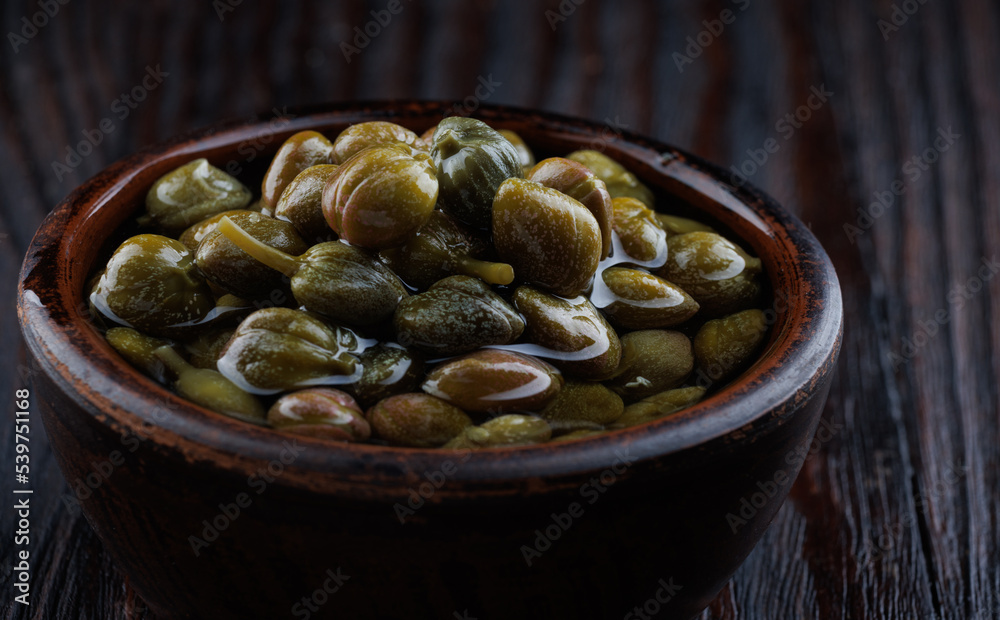 An earthenware bowl filled to the brim with capers stands on a wooden table. An earthenware bowl filled to the brim with capers stands on a wooden table.