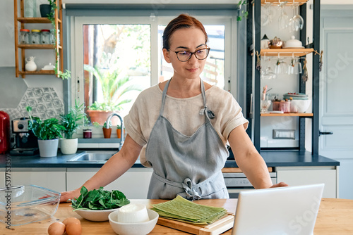 Woman preparing homemade lasagna with spinach and ricotta and looking for recipe online in her laptop. Traditional italian dish. Selective focus.