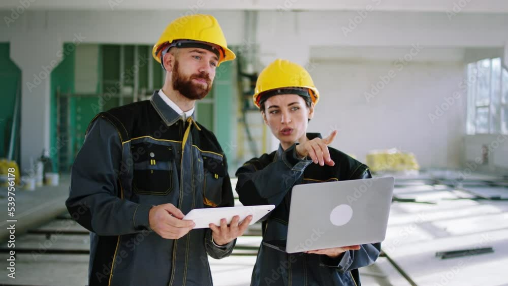 In front of the camera good looking woman engineer and her assistant walking through the large space building woman holding a laptop and the man holding a digital tablet to analysing the plan of In front of the camera good looking woman engineer and her assistant walking through the large space building woman holding a laptop and the man holding a digital tablet to analysing the plan of