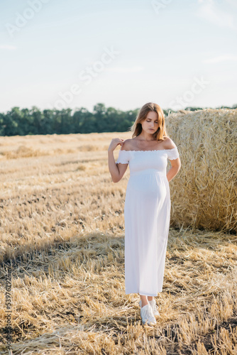 beautiful pregnant brunette in a white dress near the haystacks in the field at sunset. High quality photo