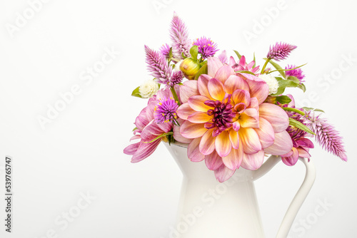 Bouquet of a variety of dahlias, celosia, and strawflower flowers. Arrangement is placed in a white vessel with a handle. White background. Space for text.