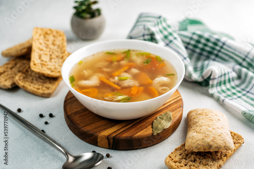 
chicken soup broth with vegetables in a white bowl, next to a spoon, bread, towel, spices
