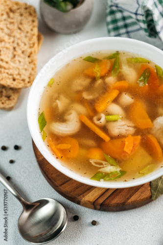  chicken soup broth with vegetables in a white bowl, next to a spoon, bread, towel, spices