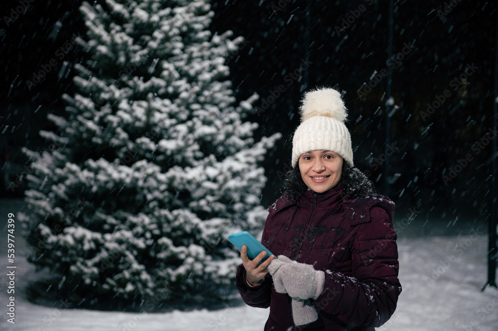 Beautiful Latin American woman in warm clothes, wool mittens and hat, holding smartphone and smiling a toothy smile looking at camera, standing against a tall fir-tree in the snow covered city street Beautiful Latin American woman in warm clothes, wool mittens and hat, holding smartphone and smiling a toothy smile looking at camera, standing against a tall fir-tree in the snow covered city street