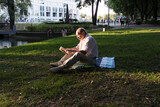 An elderly man in a white shirt is sitting on a blanket, on the ground in a park and reading an interesting book. A pensioner alone is resting in nature, passionate about his hobby.