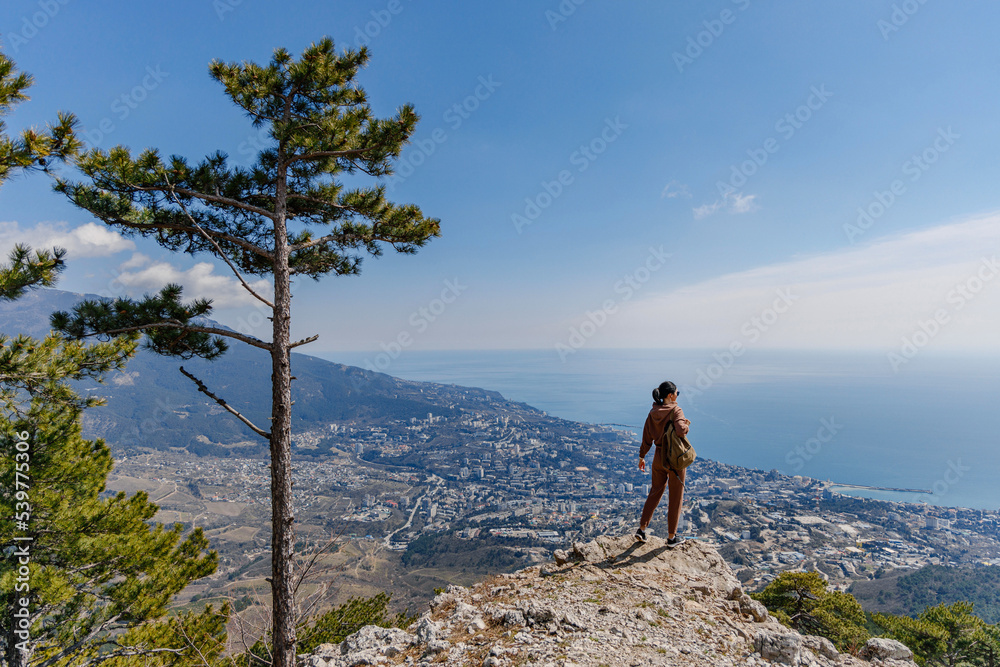 A happy woman stands on top of a mountain and looks at the sea and the city in the distance