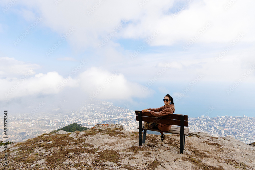 Woman sitting on a wooden bench on top of a hill with sky and city in the background on a sunny day