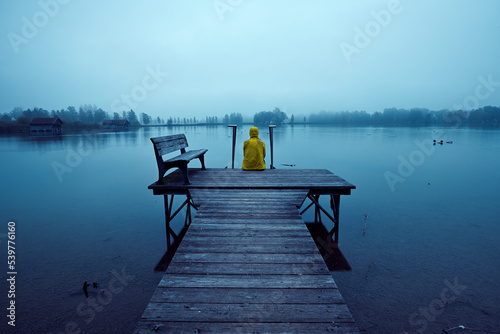 Eine Person sitzt einsam auf einem Bootssteg an einem See bei düsterer Stimmung 