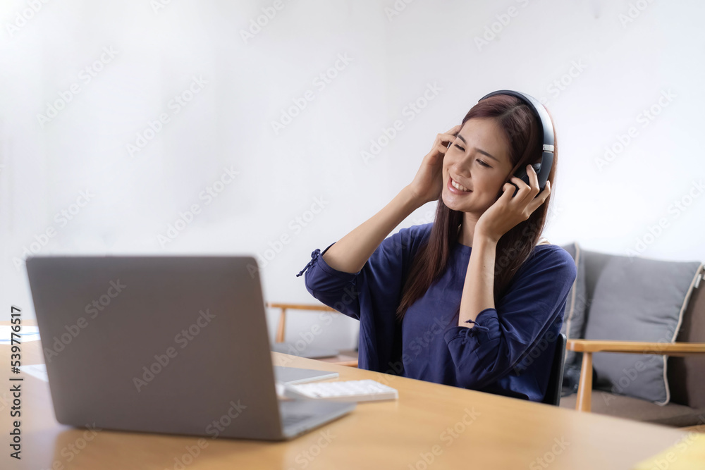 Asian woman with headphones working on a laptop and listening music at home office. WFH. Work from home. Prevention Coronavirus COVID-19 concept. Asian woman with headphones working on a laptop and listening music at home office. WFH. Work from home. Prevention Coronavirus COVID-19 concept.