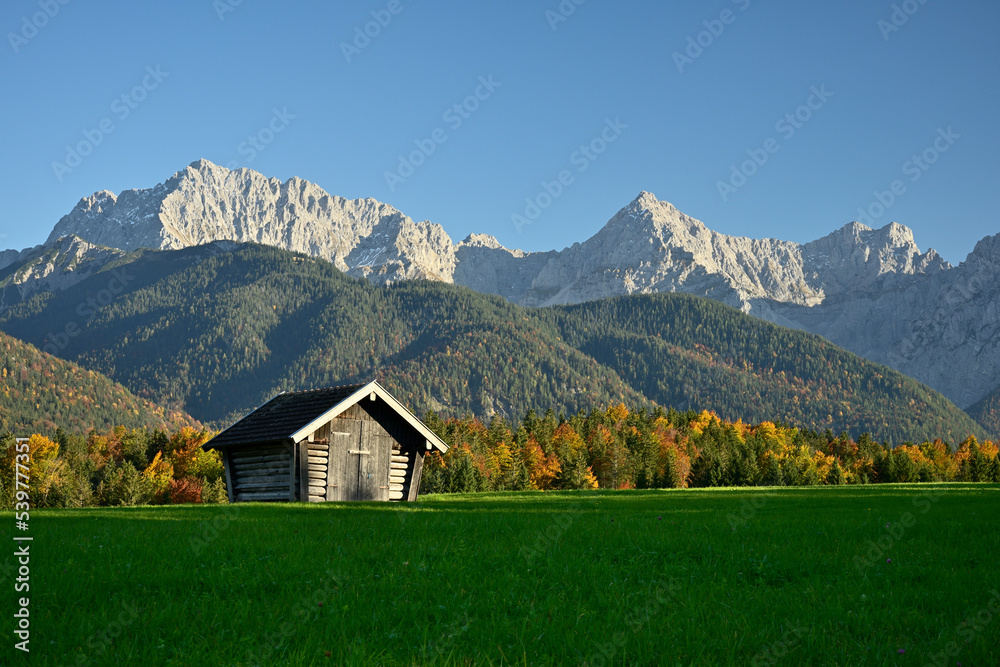 Ein Heuschober steht auf einer grünen Wiese vor einem herbstlich bunten Wald und dem Karwendel Gebirge bei blauem Himmel.