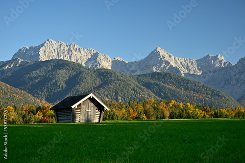 Ein Heuschober steht auf einer grünen Wiese vor einem herbstlich bunten Wald und dem Karwendel Gebirge bei blauem Himmel.