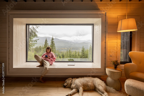 Woman sits with mobile phone on window with great view on mountains behind while resting with her dog in house on nature. Recreation and escaping to nature concept