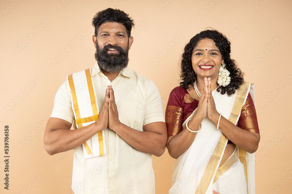 Happy South indian couple wearing traditional white dress standing in a greeting pose to Namaste hands isolated on beige background, man wearing lungi and woman wearing saree. Happy South indian couple wearing traditional white dress standing in a greeting pose to Namaste hands isolated on beige background, man wearing lungi and woman wearing saree.