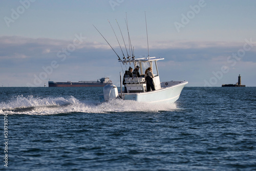 Center console fishing boat heading toward a lighthouse with a ship in the background.