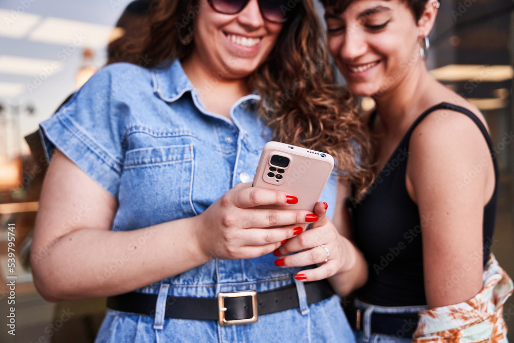 Cheerful girlfriends using smartphone near glass wall Cheerful girlfriends using smartphone near glass wall