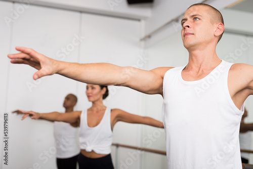 Group of people doing ballet exercises using barre in gym with focus to fit athletic toned man