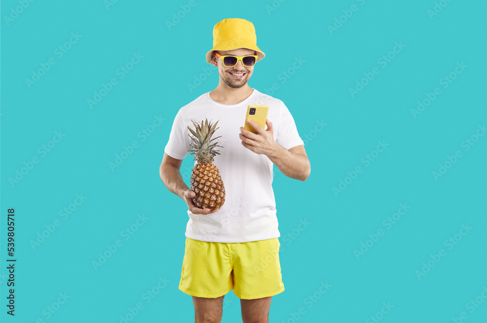 Happy man uses his mobile phone to order services, pay online or communicate with friends during summer vacation. Man in summer clothes with pineapple and with mobile phone on light blue background. Happy man uses his mobile phone to order services, pay online or communicate with friends during summer vacation. Man in summer clothes with pineapple and with mobile phone on light blue background.