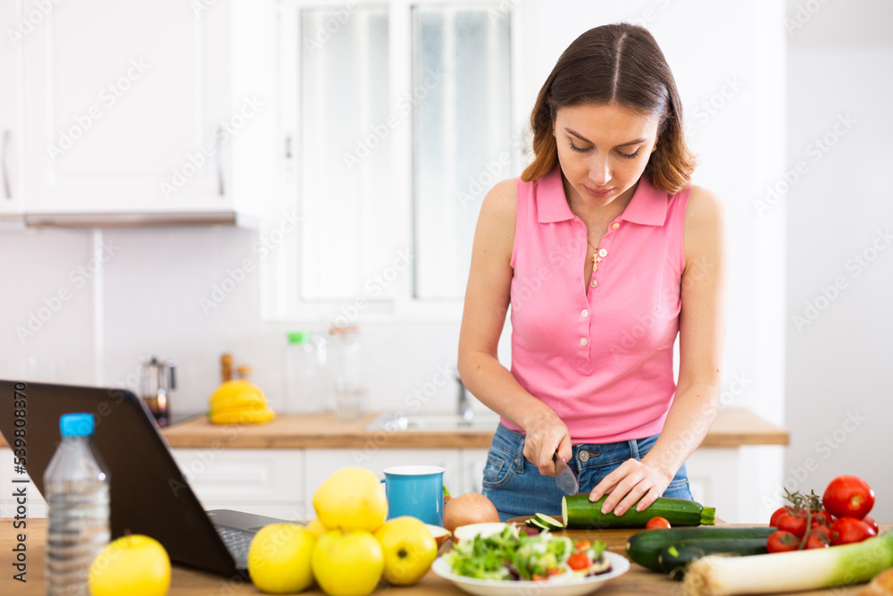 Young woman cooking vegetables for dinner and browsing sns on laptop