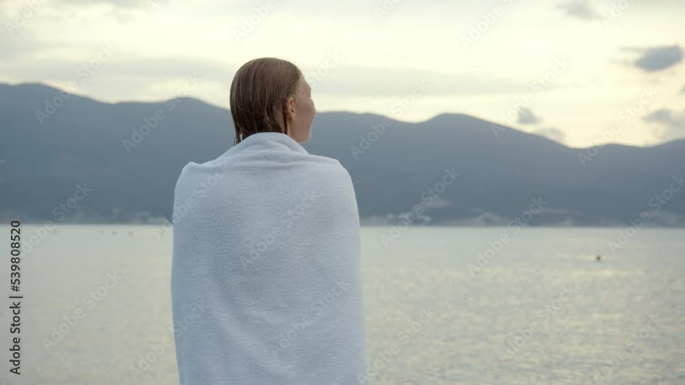 sexy slim young woman in purple swimsuit wraps herself in white towel on seashore in morning or evening. Wet girl after bathing tries to keep warm and throws towel over herself. sexy slim young woman in purple swimsuit wraps herself in white towel on seashore in morning or evening. Wet girl after bathing tries to keep warm and throws towel over herself.
