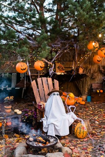 Little white halloween ghost in hat and glasses sits on wooden chair  by the fire in front of a pine tree with jack-o-lanternsHalloween celebration holiday.  Halloween consept.