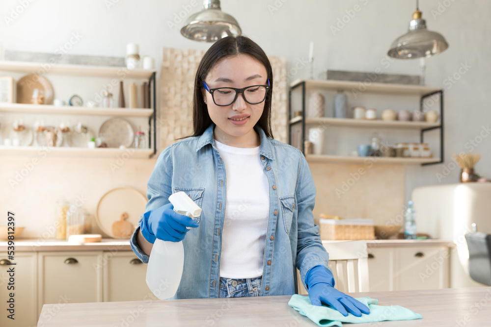 Maid services. Young beautiful Asian woman works as a cleaner. Cleans the house. Wipes the table with a cloth in rubber gloves, sprays the surface with a spray. Maid services. Young beautiful Asian woman works as a cleaner. Cleans the house. Wipes the table with a cloth in rubber gloves, sprays the surface with a spray.